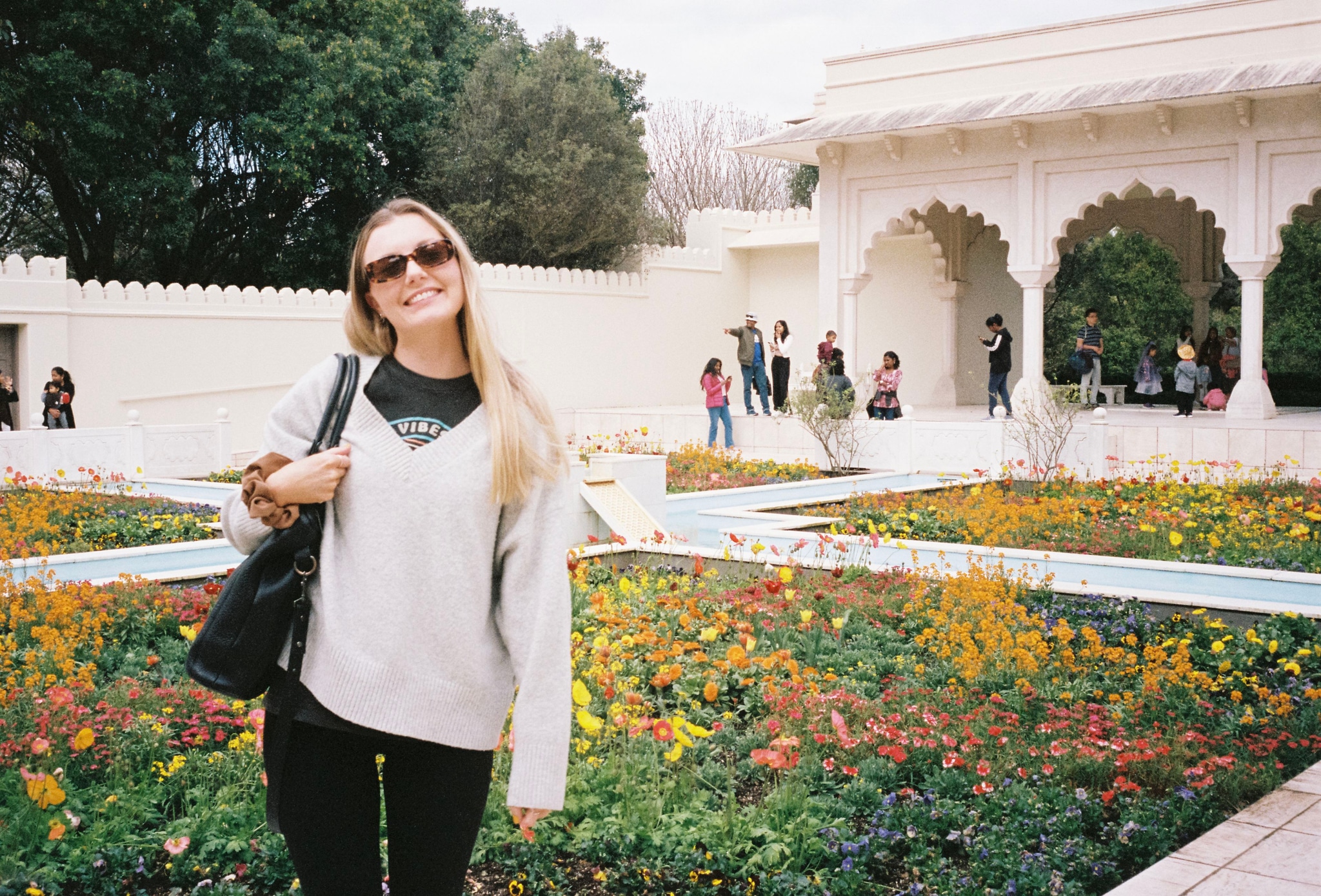 woman sunglasses sweater garden flowers building arches trees people path white green yellow black bag smile outdoors architecture