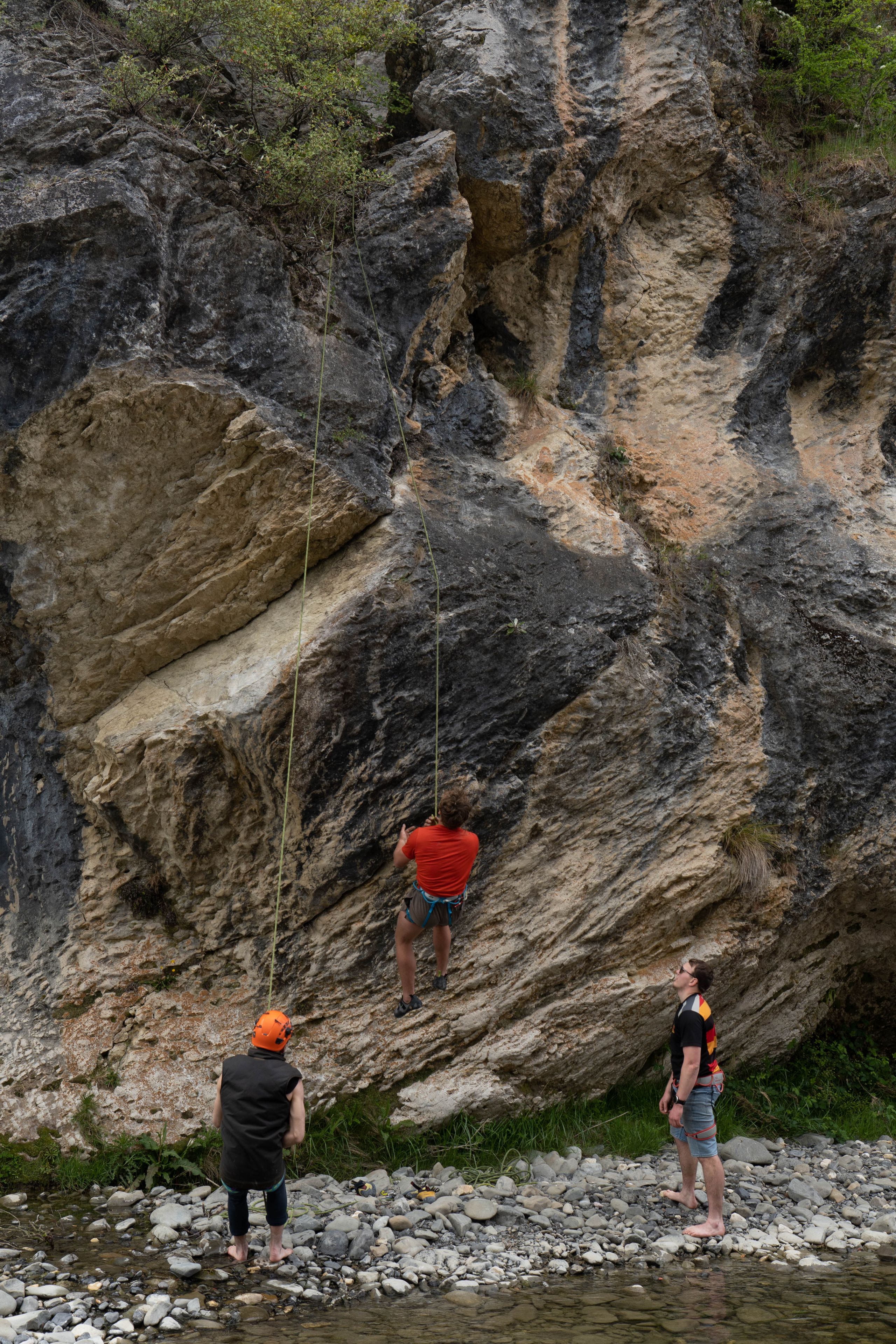 rock climbing cliff rope climber people helmet harness outdoor adventure nature stone gray red black green water pebbles safety gear teamwork sport challenge vertical ascent rugged landscape exploration activity recreation exercise physical fitness teamwork concentration balance skill determination primary color gray