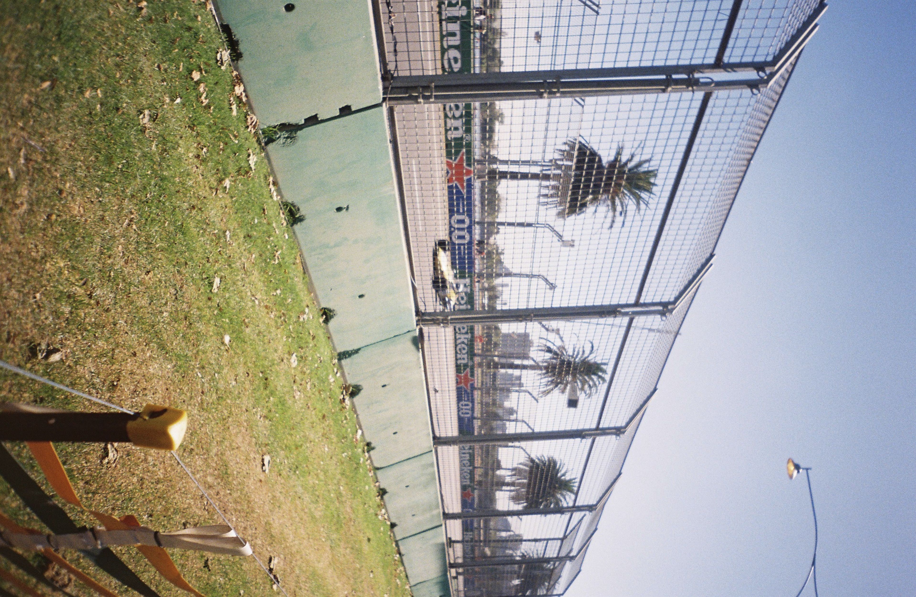 racecar track fence palm trees grass sky blue road barrier sunlight shadow pole lamp post advertising sign leaves pavement