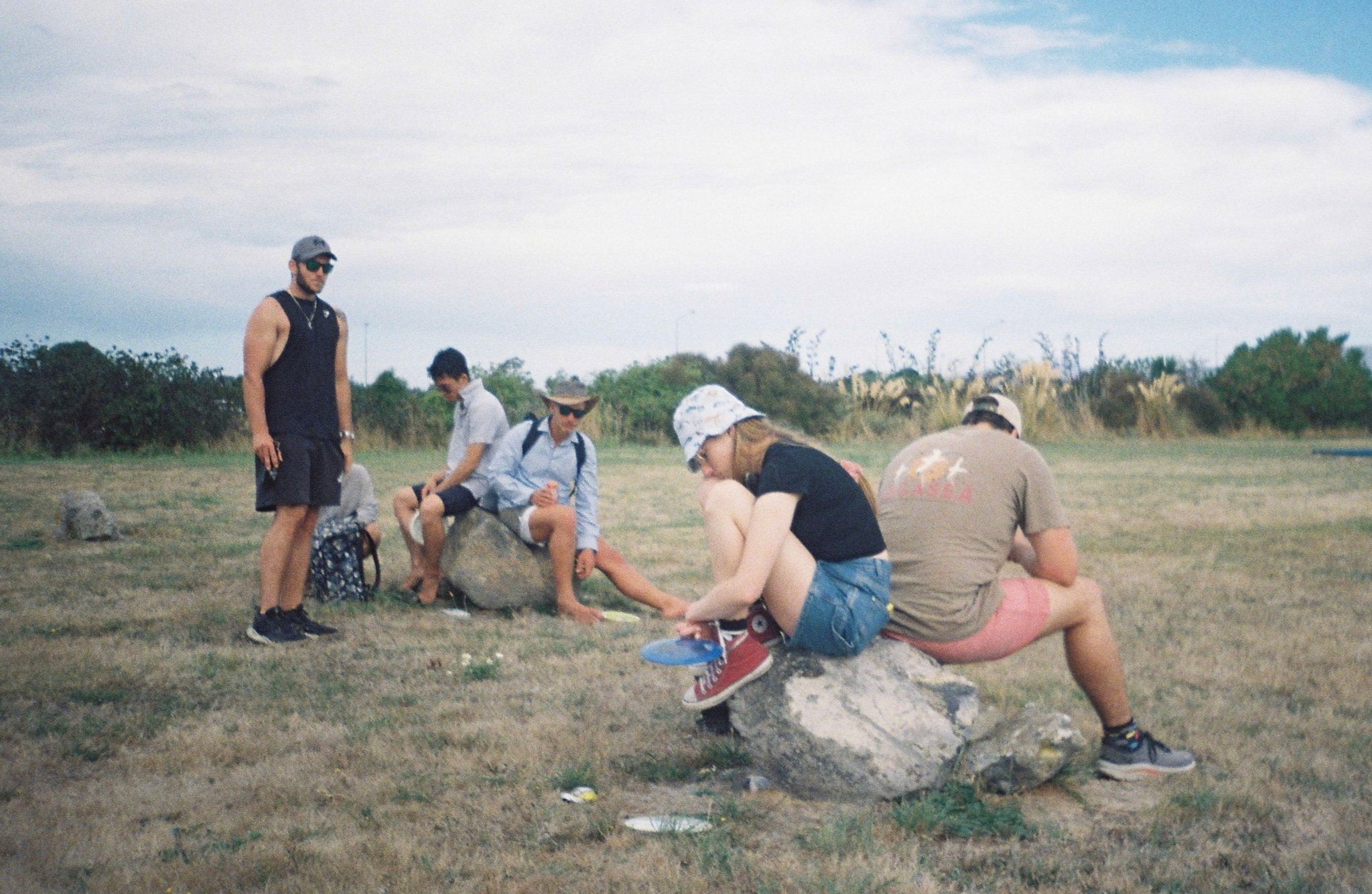 people outdoors grass sky rocks sitting standing hats shoes shorts t-shirts clouds trees field nature casual gathering group summer leisure activity landscape blue green brown gray white relaxed conversation social interaction day light natural environment open space rural scene recreation friendship companionship exploration adventure travel journey relaxation tranquility peacefulness harmony unity connection bonding enjoyment fun freedom simplicity minimalism serenity calmness balance perspective distance horizon openness vastness expanse