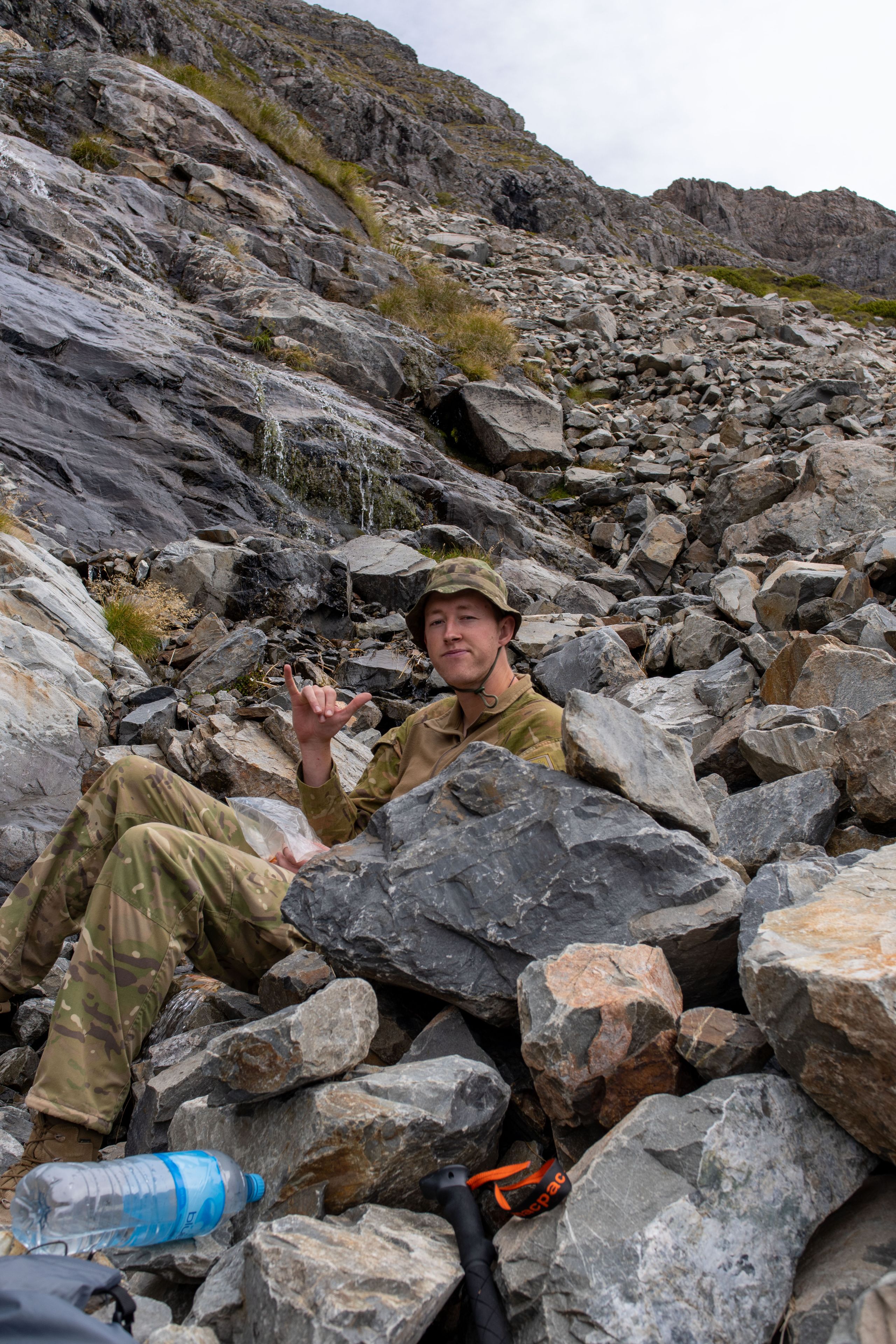 rocks camouflage clothing hat water bottle backpack mountains outdoor hiking sitting gray brown green sky rugged terrain