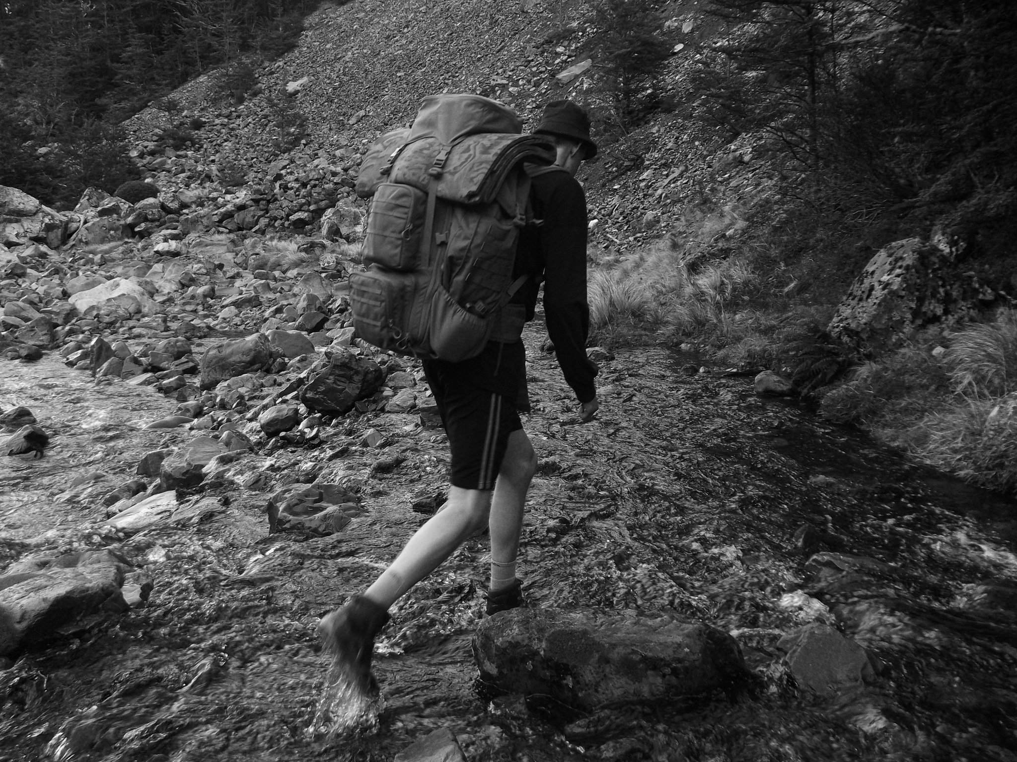 hiking backpack river rocks forest outdoors nature stream crossing boots black white grayscale trekking adventure wilderness exploration journey rugged landscape water trees trail