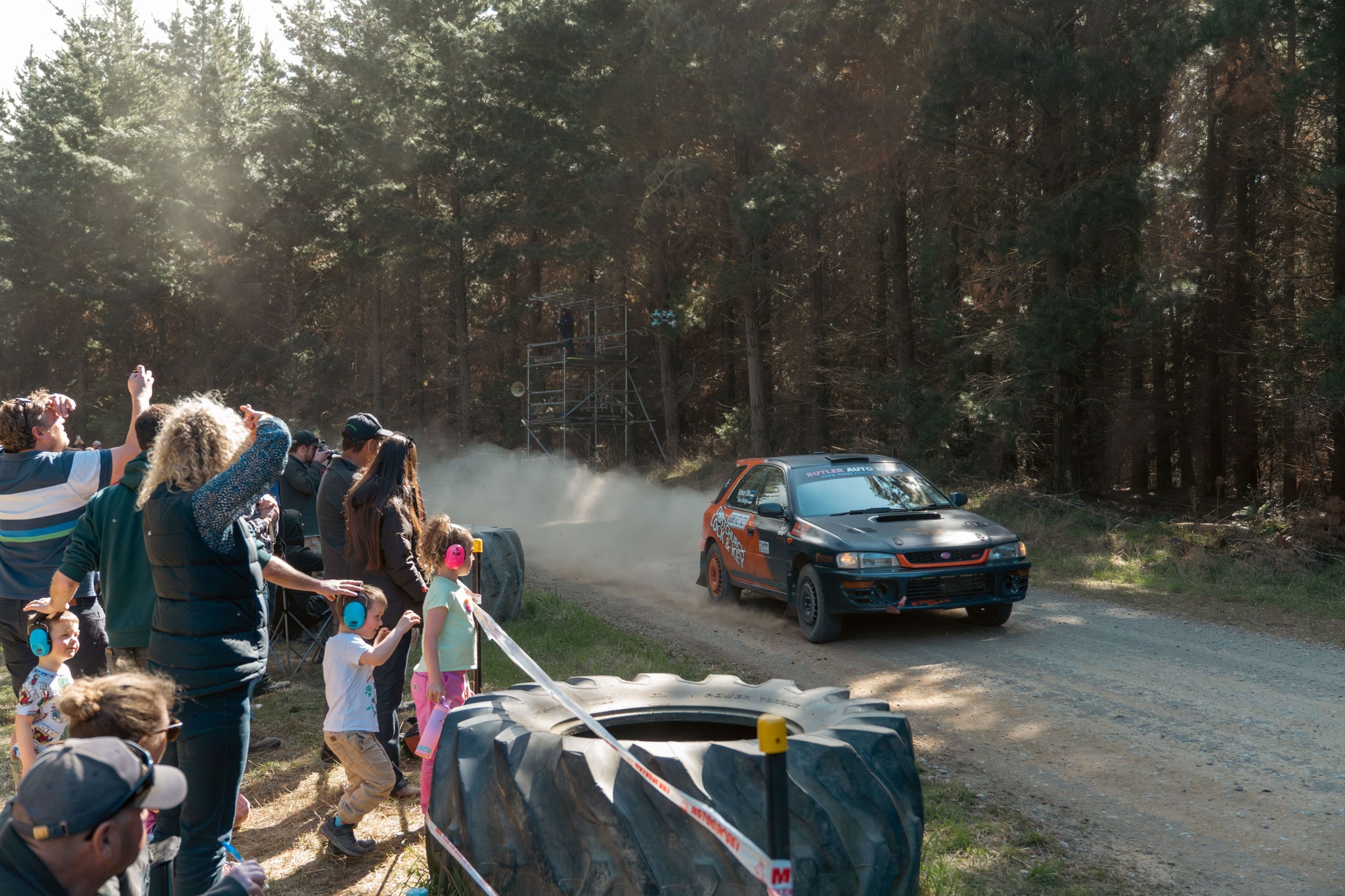 car race rally dirt road spectators trees forest dust motion sunlight blue green red black white gray people children adults fence cheering action speed excitement outdoors daytime