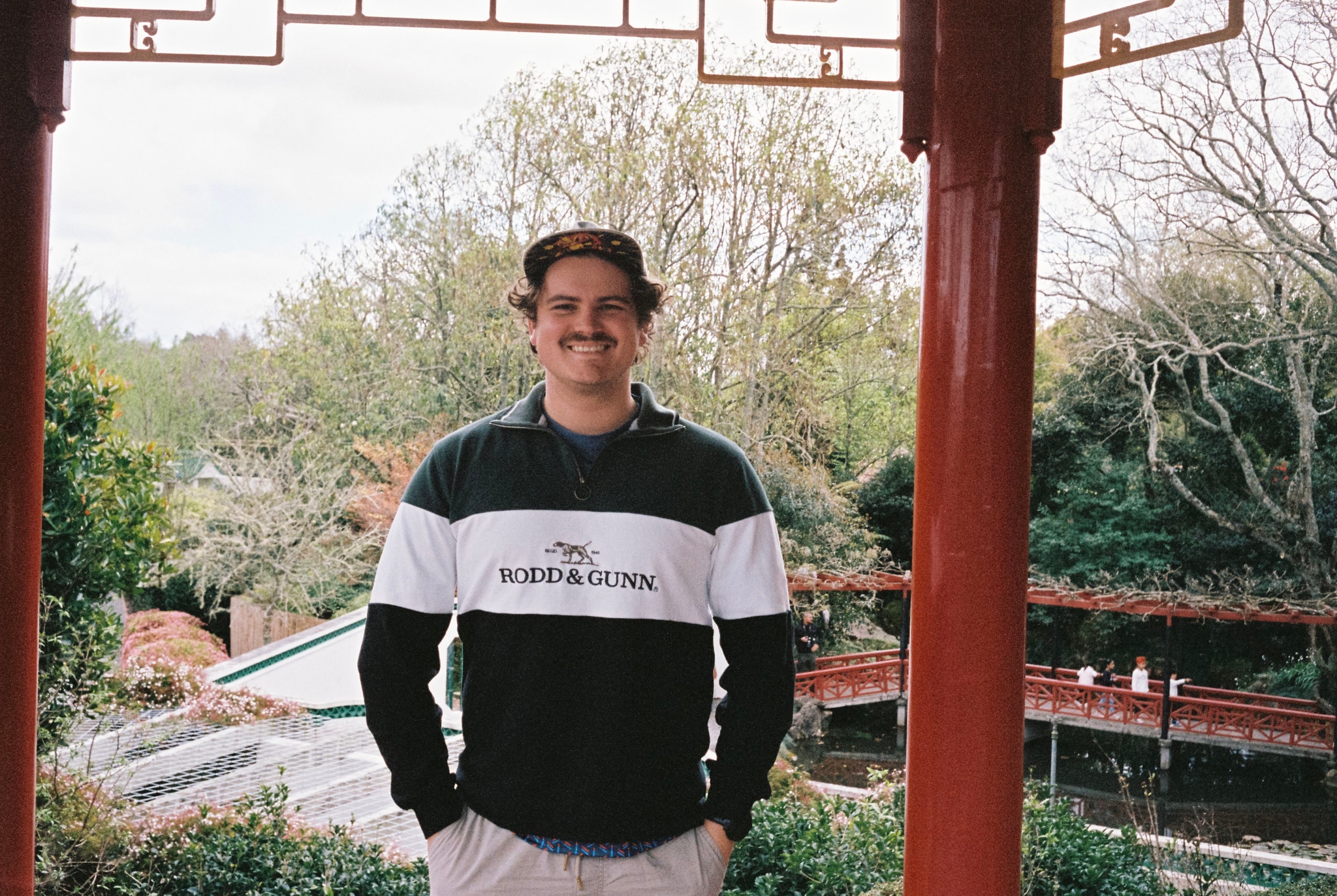 person man smiling standing outdoors trees greenery garden pavilion red columns roof sky casual clothing hat mustache nature architecture landscape background spring foliage leisure portrait photography travel tourism relaxation peaceful serene primary color red