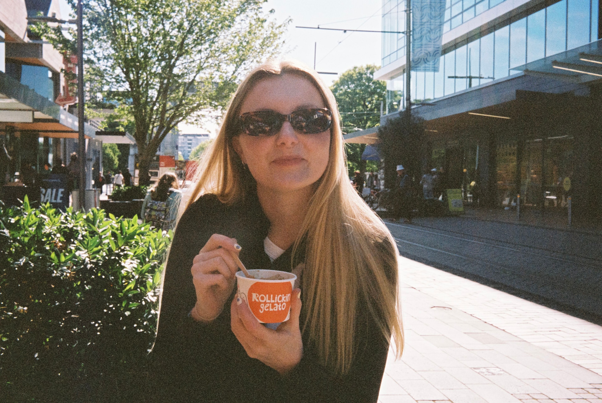 woman sunglasses ice cream spoon cup outdoor street building tree plants sunlight shadow casual clothing blonde hair long hair urban scene daytime primary color green