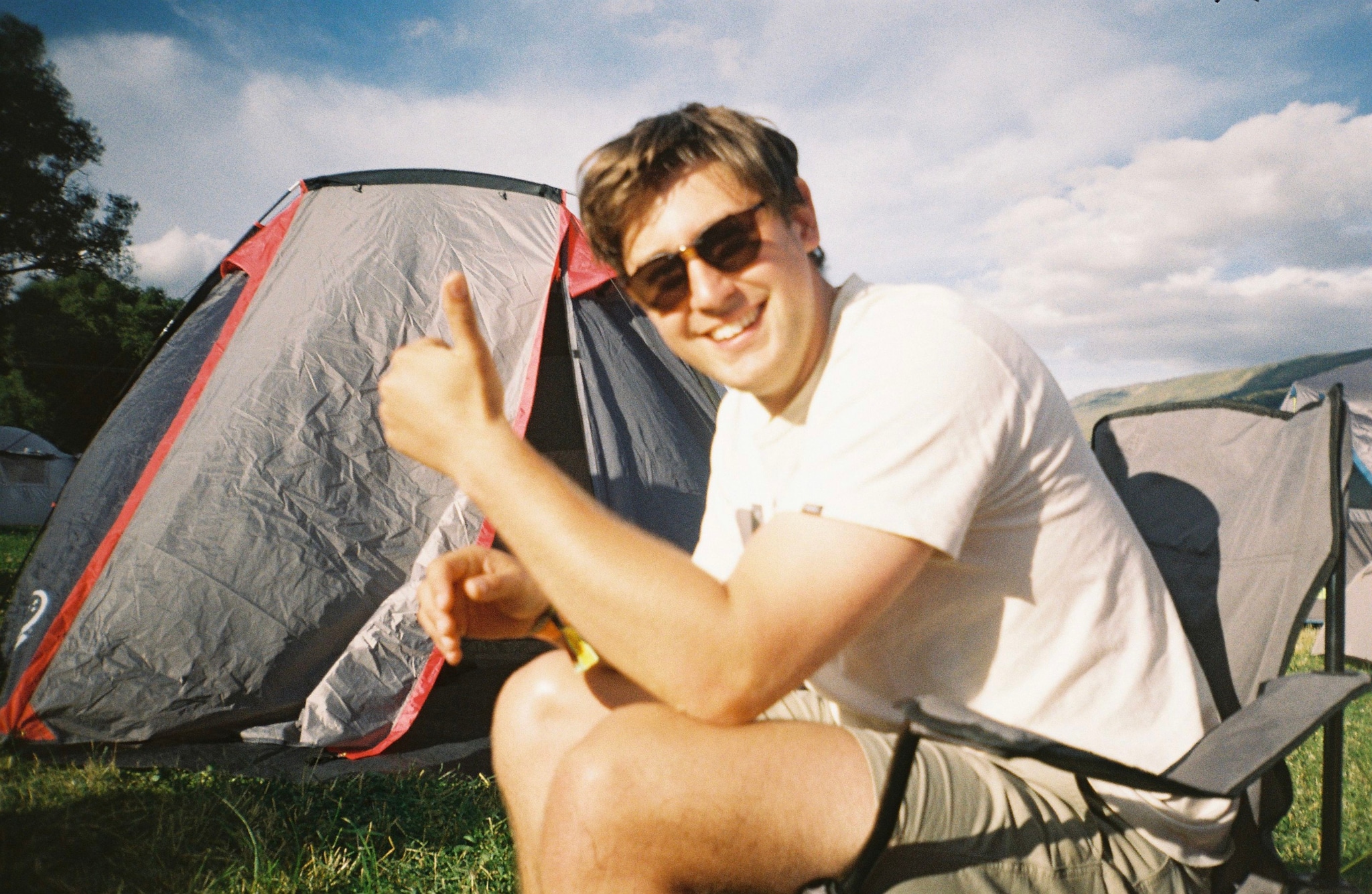 Man Tent Sunglasses Chair Grass Sky Cloud Thumbs Up Bottle Camping Outdoor Daylight Casual Relaxation Smiling Summer Leisure Nature Travel Adventure Recreation Happy Fun Shade Sunlight Blue Green Red White Black