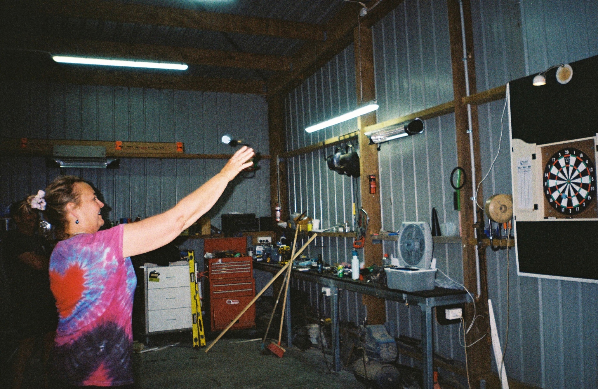 person dartboard darts throw tie-dye shirt heart pattern workshop tools shelves broom light bulb fluorescent light metal wall dart game television monitor cables wires primary color blue