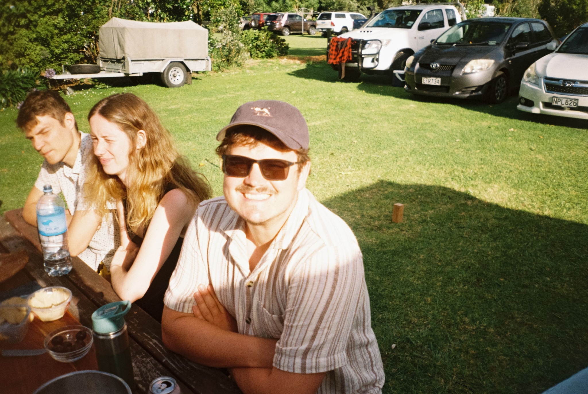 people outdoors grass cars table bottle cap sunglasses smile trees sunlight shadow blue white green brown black red gray