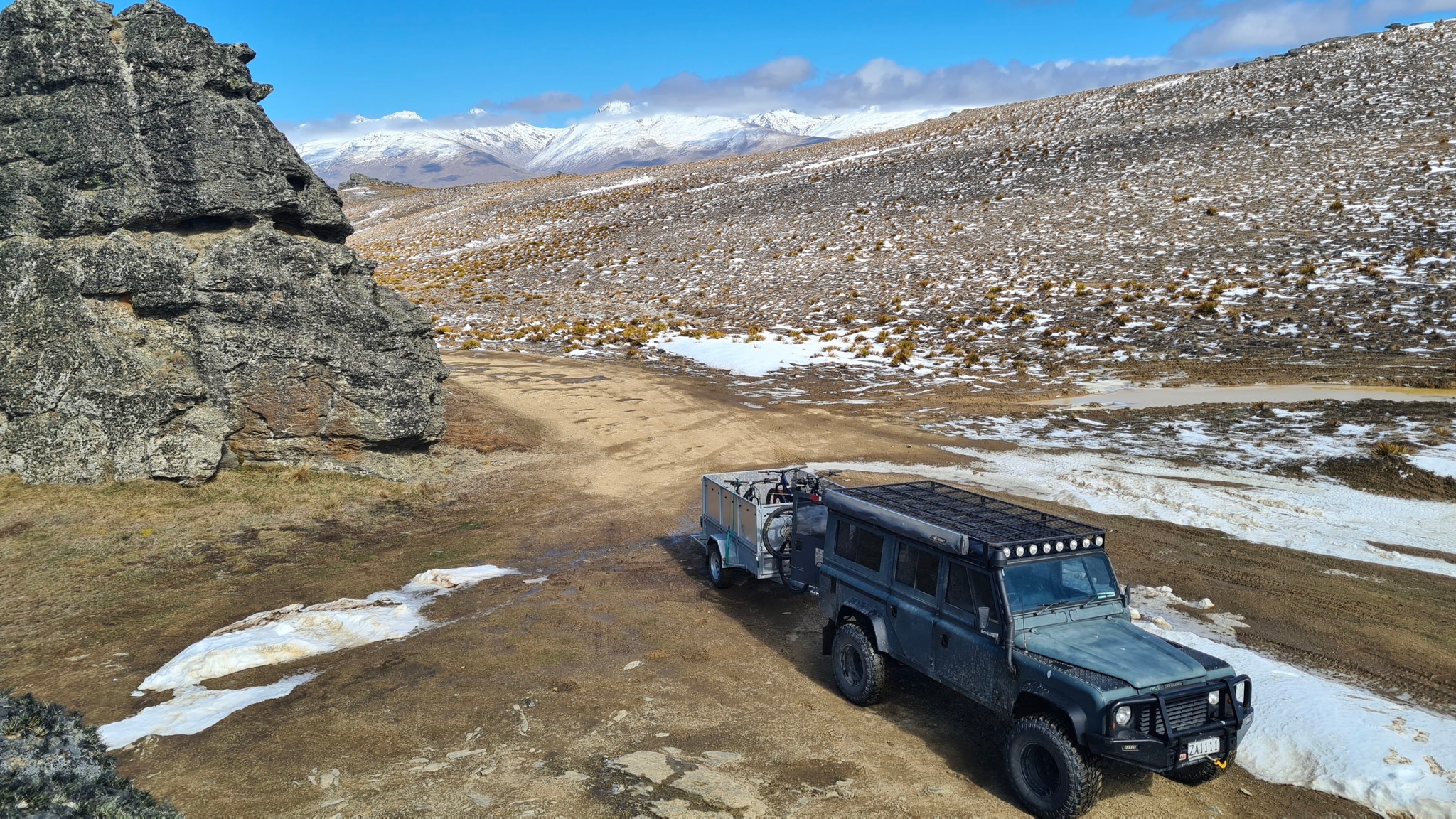 A rugged off-road vehicle with a trailer is parked on a rocky terrain with patches of snow, surrounded by mountains under a clear blue sky