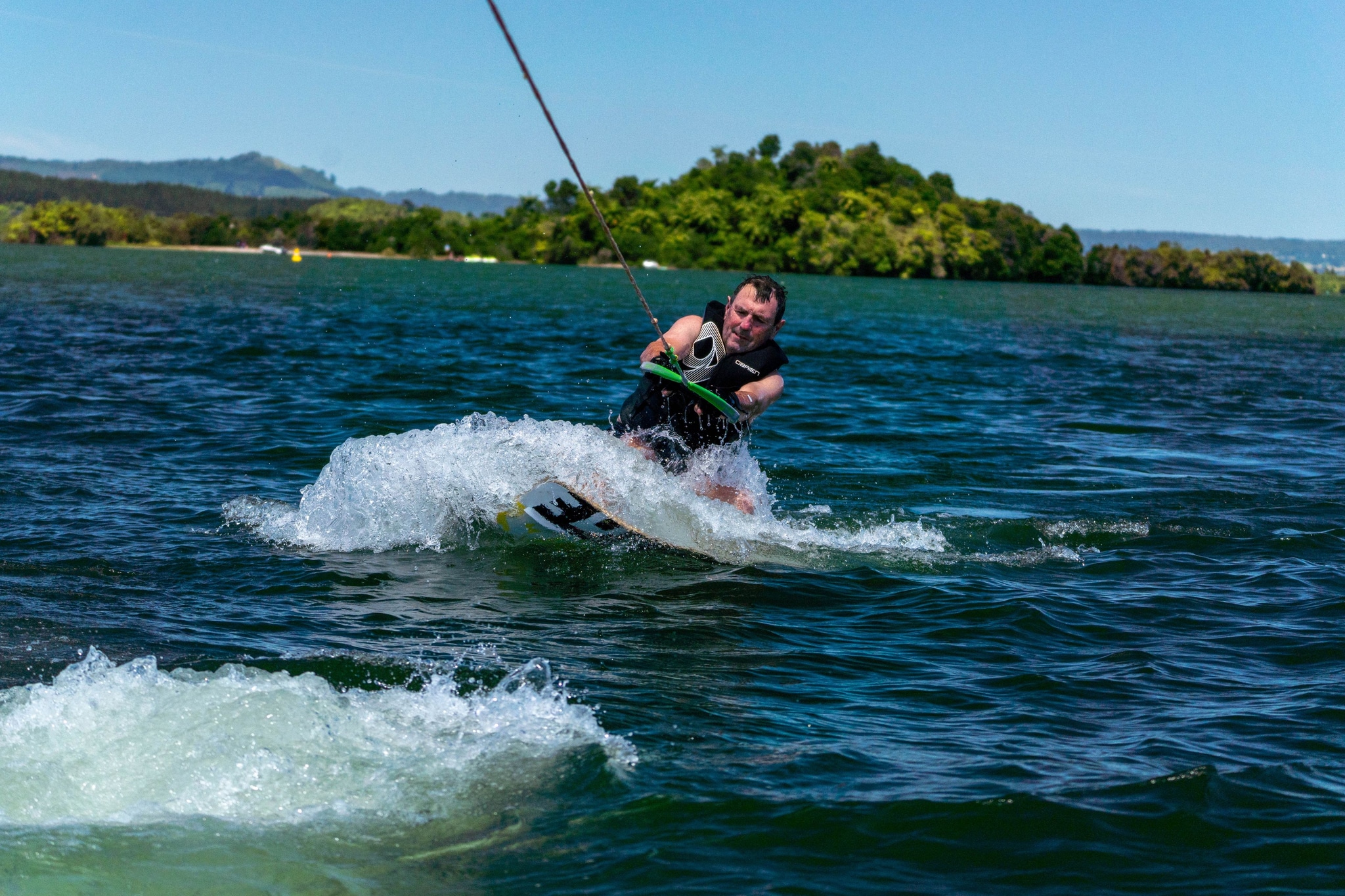 water wakeboarding wakeskate person rope waves splash lifejacket board lake outdoor sport action summer green blue sky island trees landscape adventure recreation fun movement speed thrill aquatic nature scenery activity excitement balance skill riding maneuvering primary-color-blue