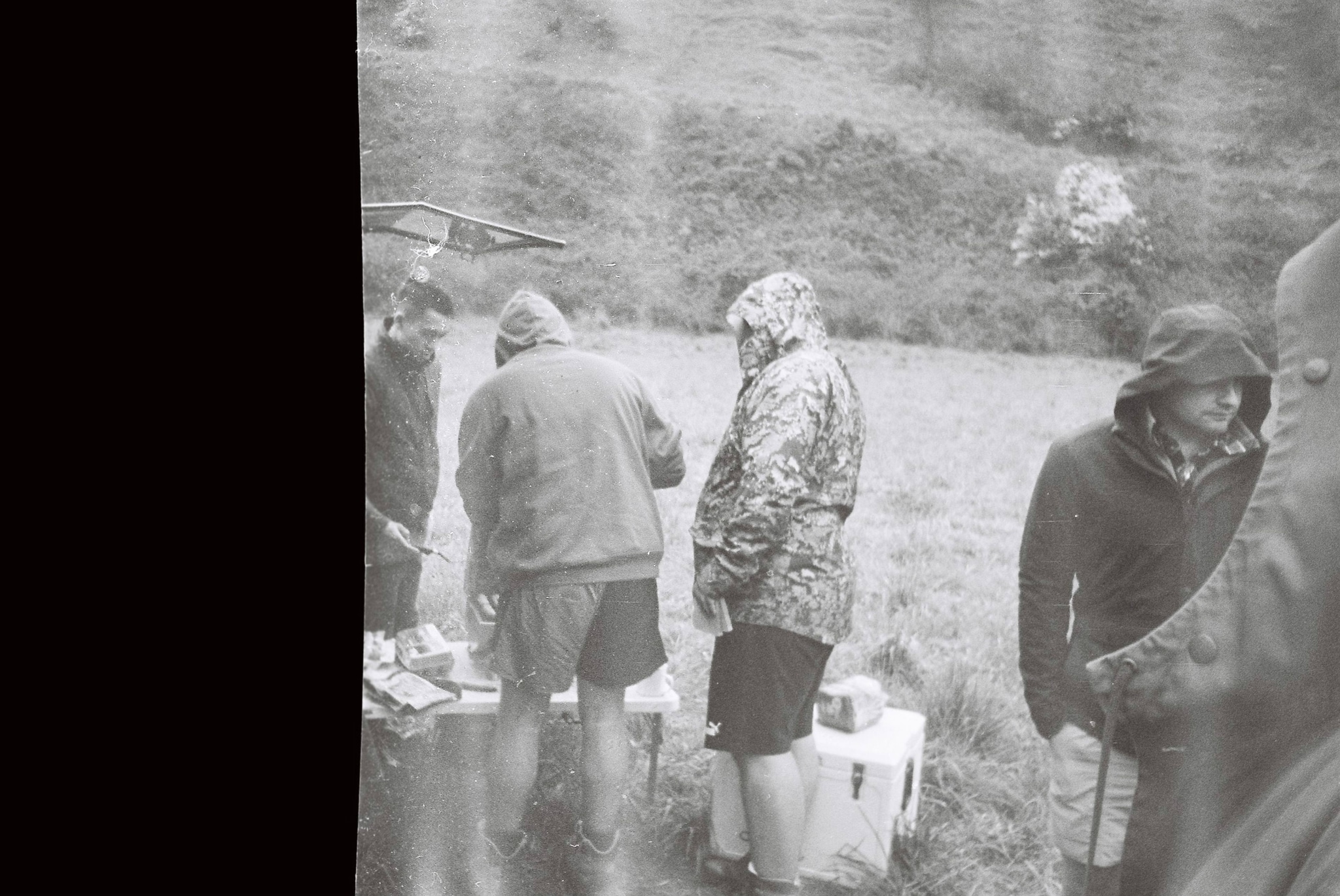 black white people outdoors coats hoods grass field table gathering overcast jackets boots nature landscape group standing rain casual clothing countryside rural scene picnic socializing weather dreary monochrome vintage retro atmosphere