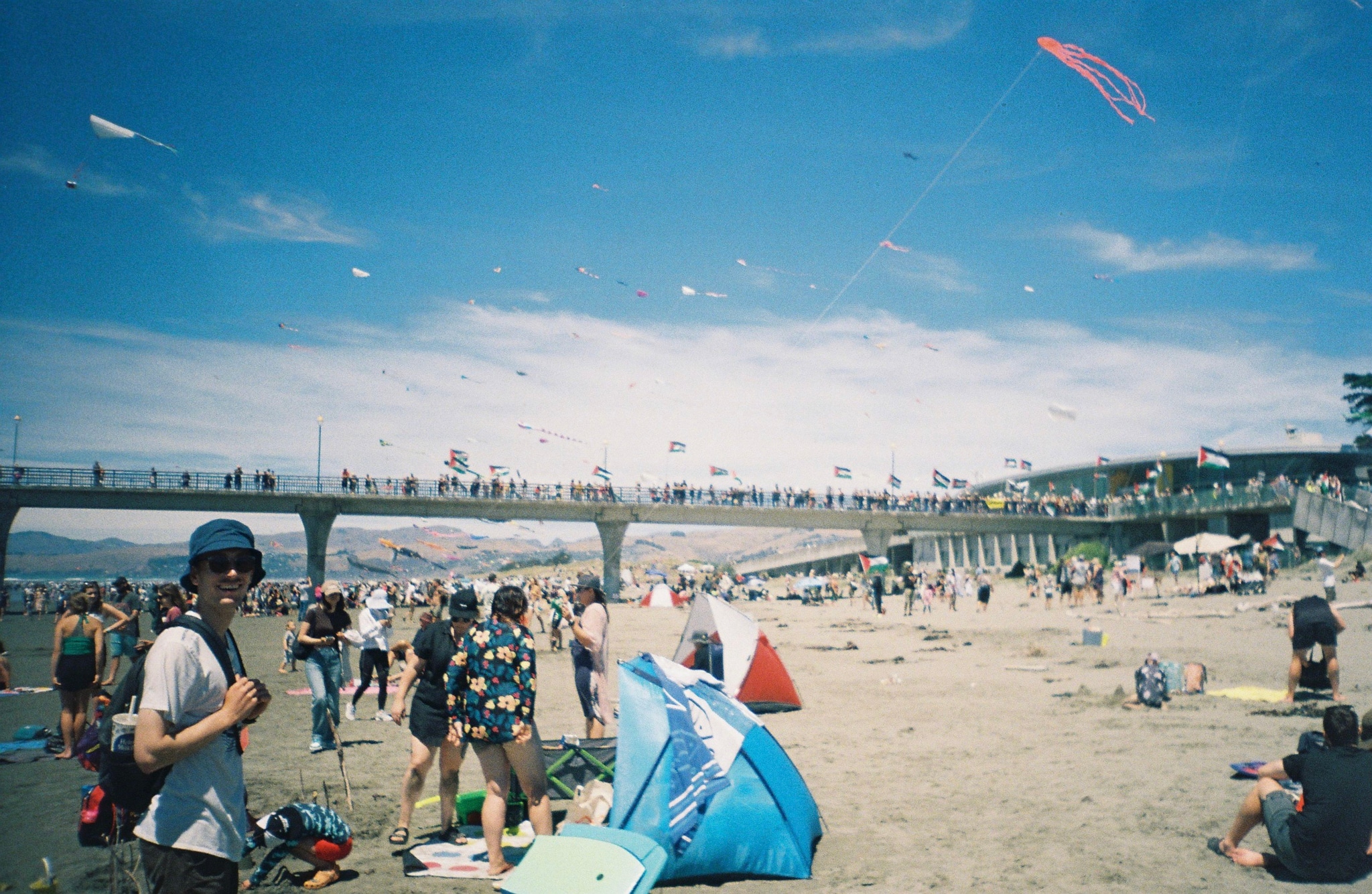 Beach Sand People Bridge Kites Sky Blue Clouds Ocean Water Sunbathers Umbrellas Towels Sunglasses Hats Swimsuits Shorts T-shirts Bags Shoes Flags Pier Summer Recreation Relaxation Fun Leisure Outdoors