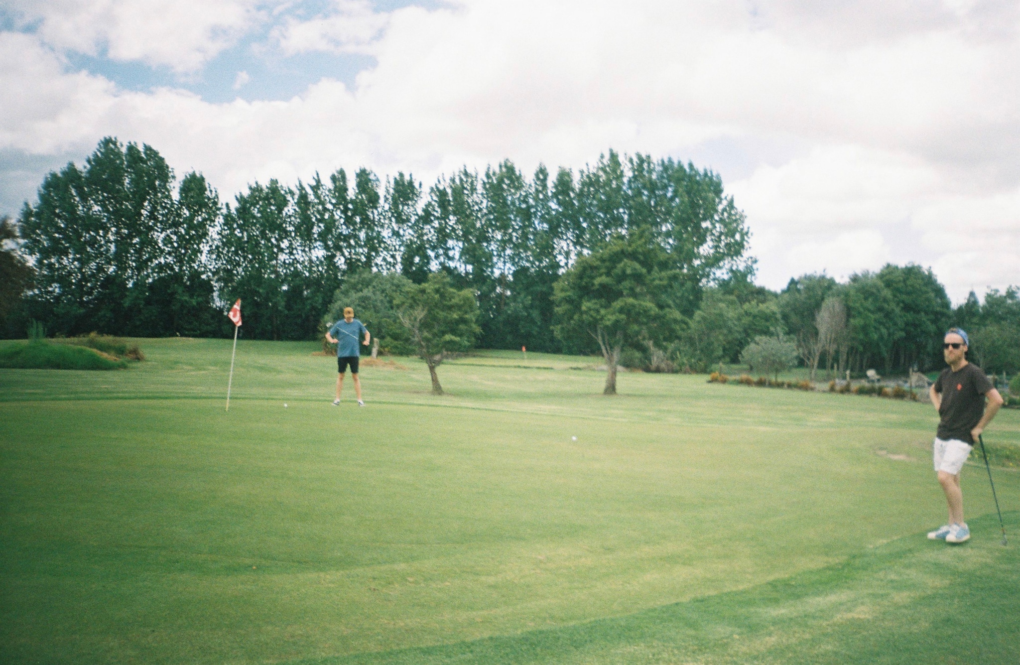 golf course grass trees sky flag pole people outdoors green blue clouds landscape sports leisure nature field fairway golfer putting hole summer day activity recreation scenery