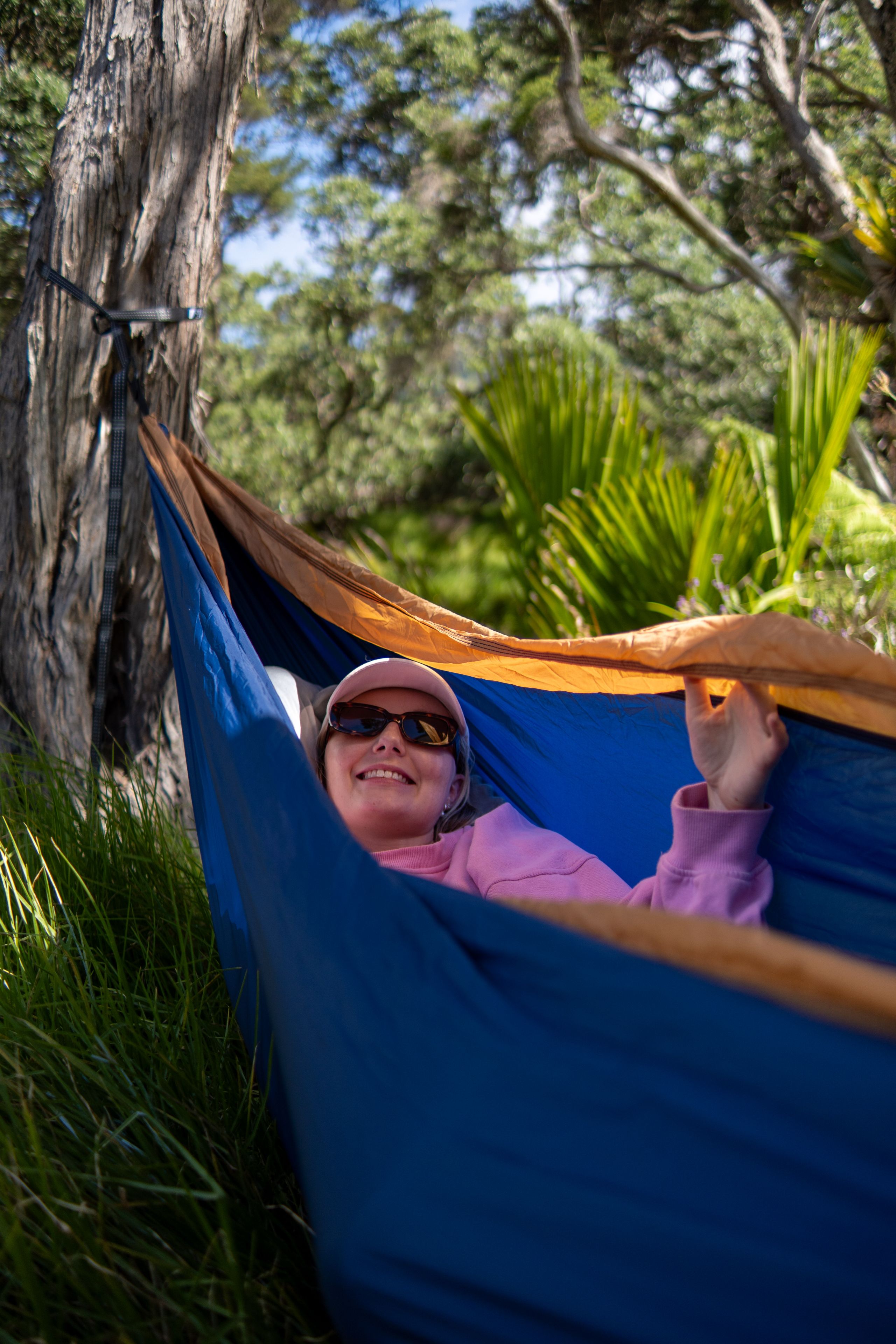 hammock tree grass person sunglasses hat smile blue pink outdoor nature relaxation forest leaves branches sunlight shade casual leisure summer