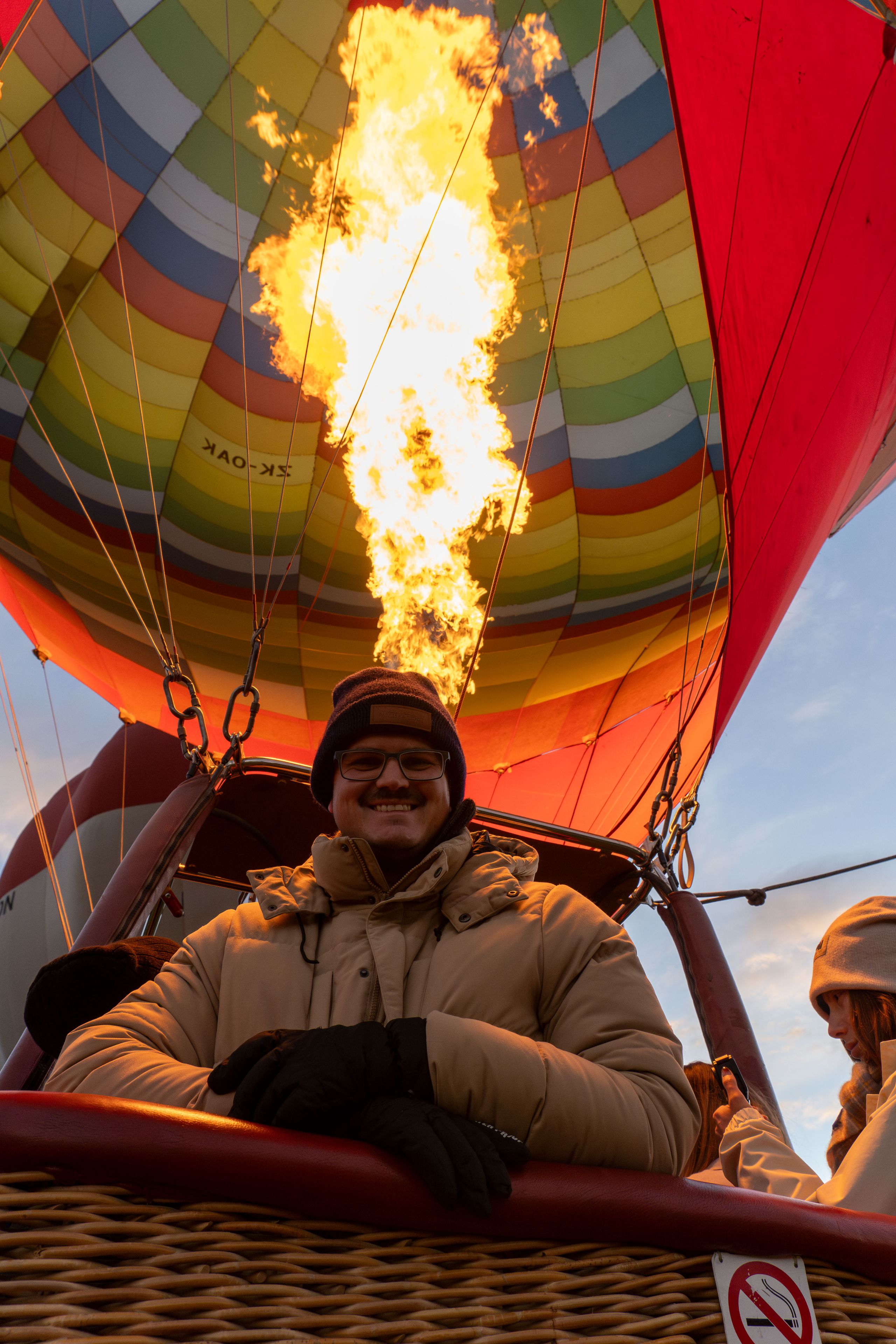 hot air balloon flame burner basket person coat gloves hat colorful sky red yellow blue green wicker ropes no smoking sign winter outdoor adventure travel flight warmth safety equipment pilot passenger vertical vertical stripes