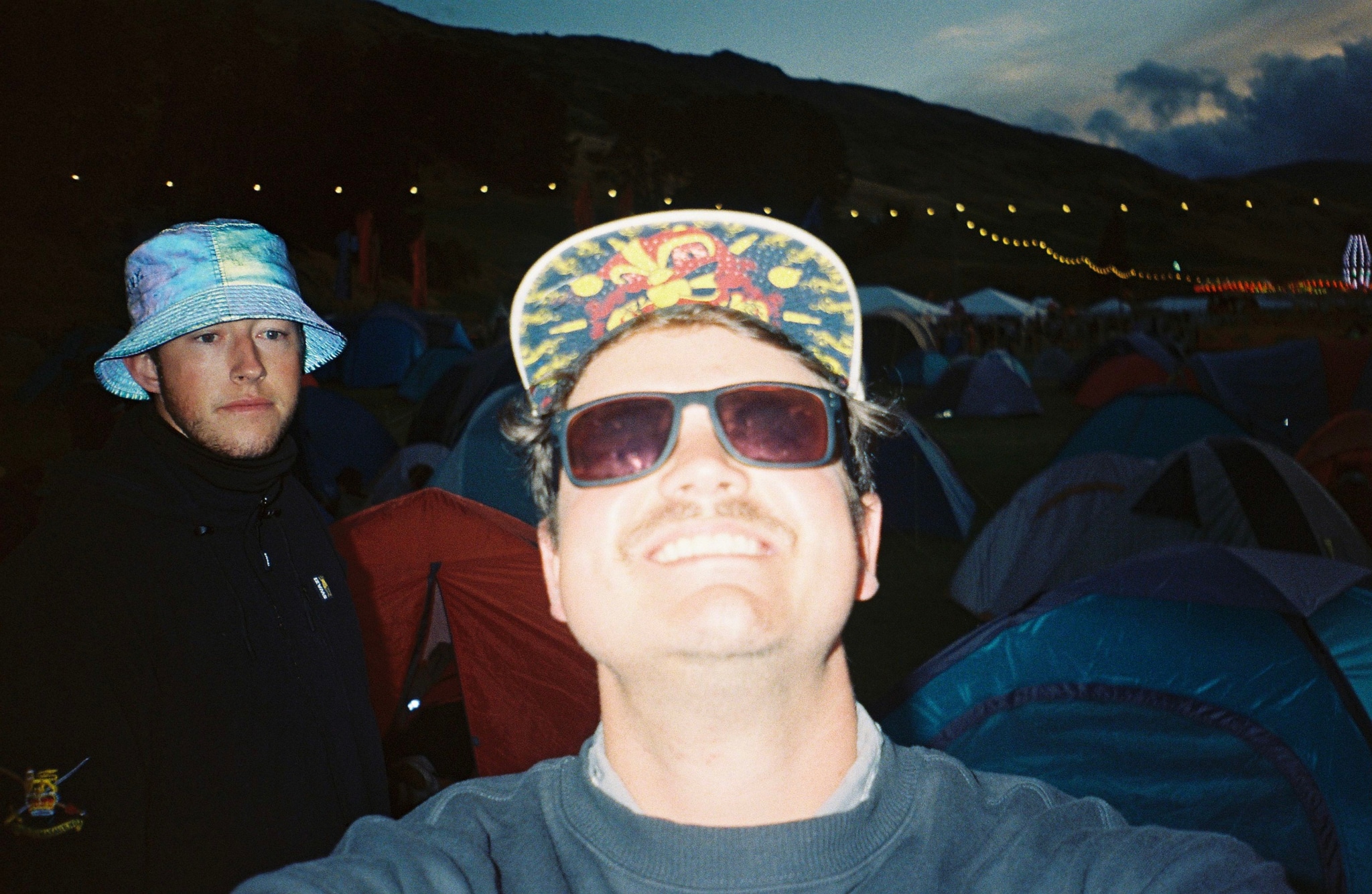 man sunglasses hat bucket hat tents camping night sky lights smiling blue red black gray festival outdoors people