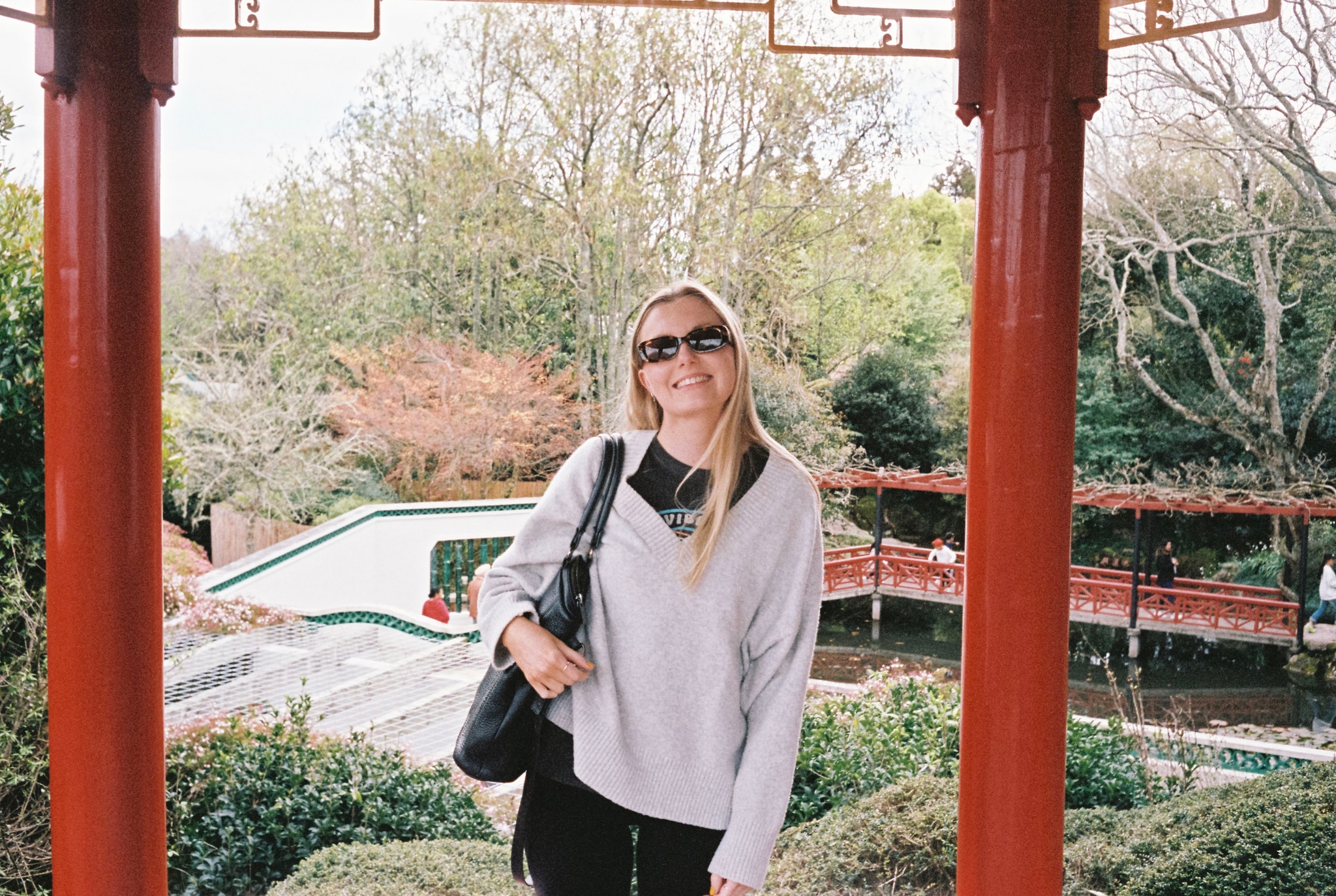 Person Woman Sunglasses Smile Blonde Hair Sweater Bag Black Red Pillars Garden Trees Bushes Pathway Bridge Outdoors Nature Daylight Sky Greenery Architecture Structure Primary Red